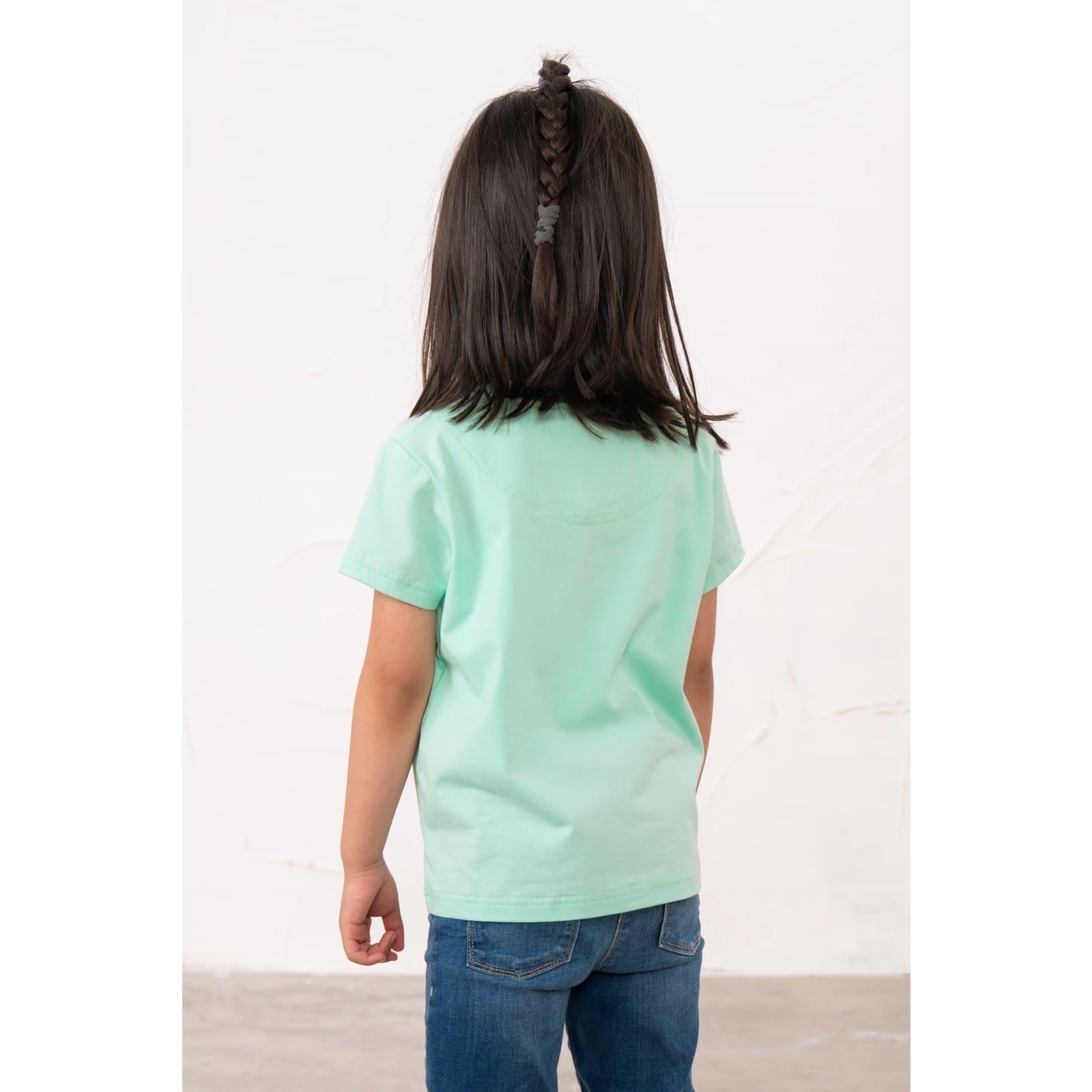 Back view of a girl wearing a kids Lighthouse short sleeve Causeway t-shirt in mint green, featuring a white and pink poodle dog print on the front.