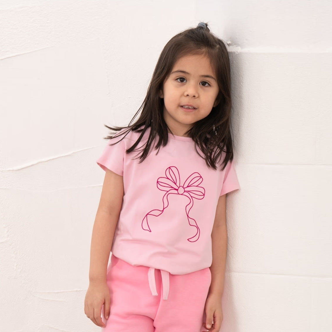 Front view of a girl wearing a Lighthouse Causeway short sleeve tee in pink with a tie hem and embroidered bow on the front.