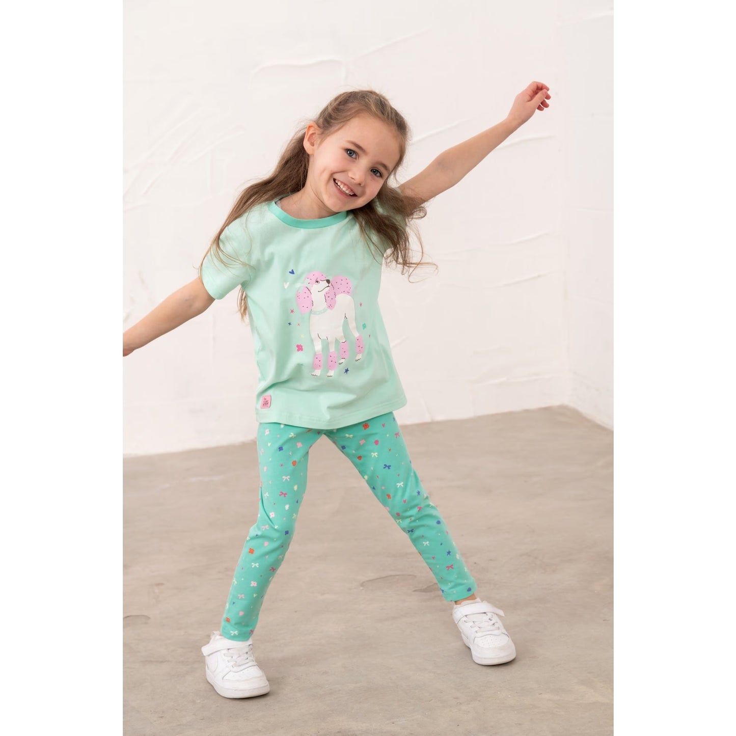 A girl wearing a pair of Lighthouse kids Mollie leggings in mint green with a bows, stars and floral pattern and a poodle print t-shirt.