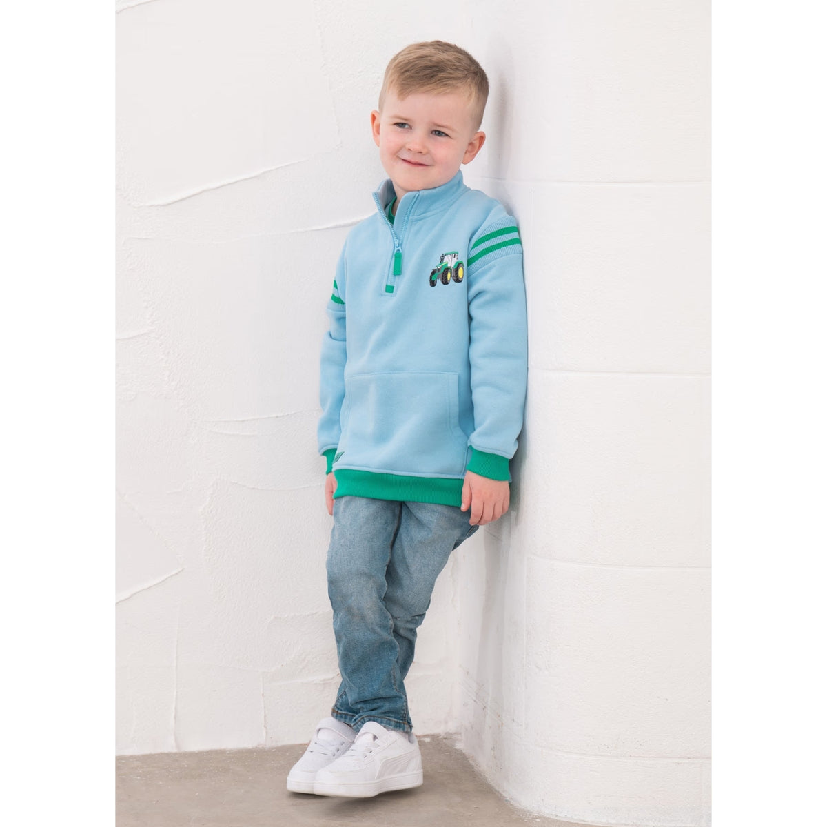 A boy wearing a Lighthouse Jonah zip neck sweatshirt in sky blue with green trim and tractor applique on the chest.
