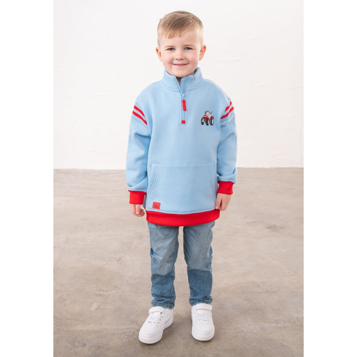 A boy wearing a Lighthouse Jonah 1/4 zip sweatshirt in sky blue with red trim and a red embroidered tractor applique on the chest.