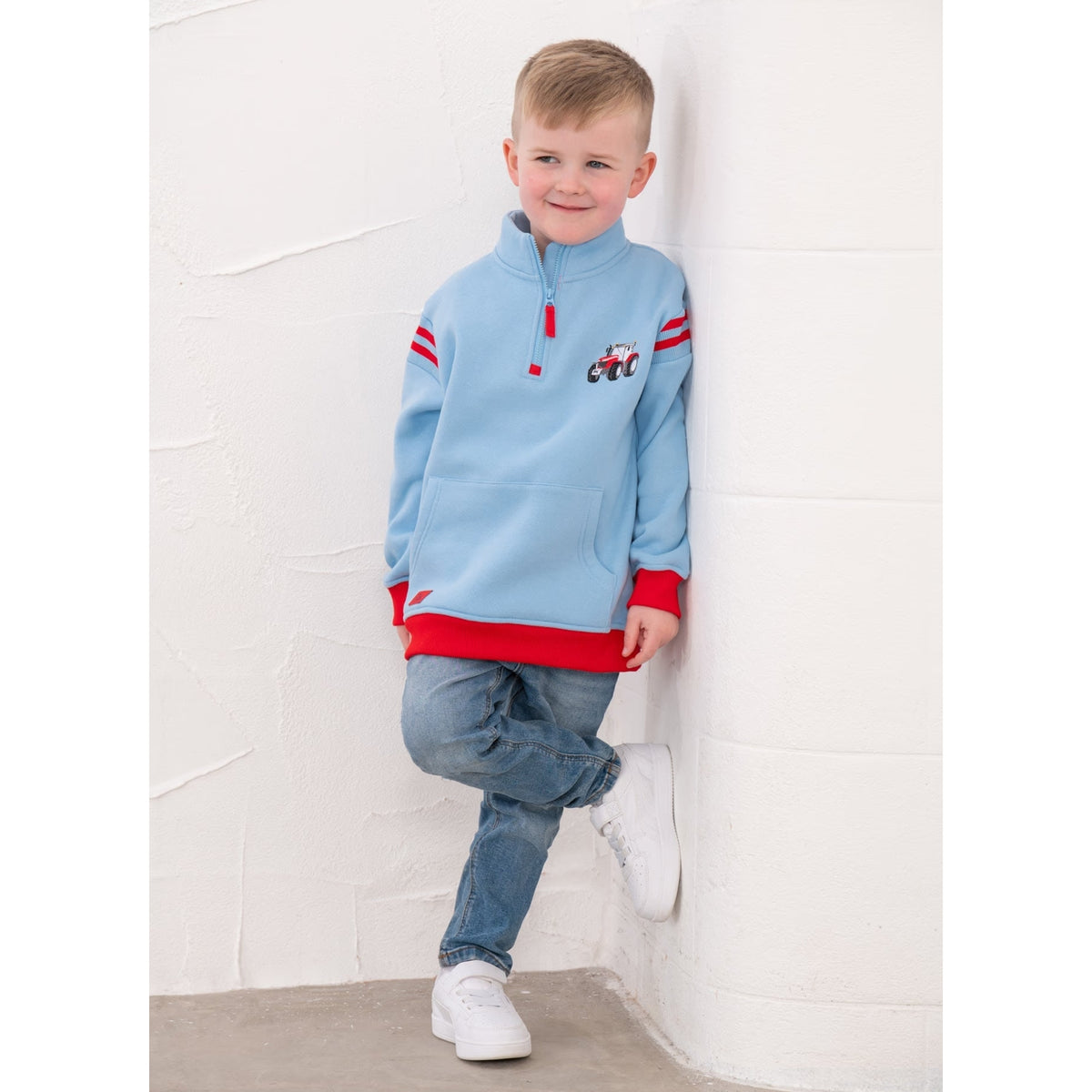 A boy wearing a Lighthouse Jonah 1/4 zip sweatshirt in sky blue with red trim and a red embroidered tractor applique on the chest with blue jeans and white trainers.