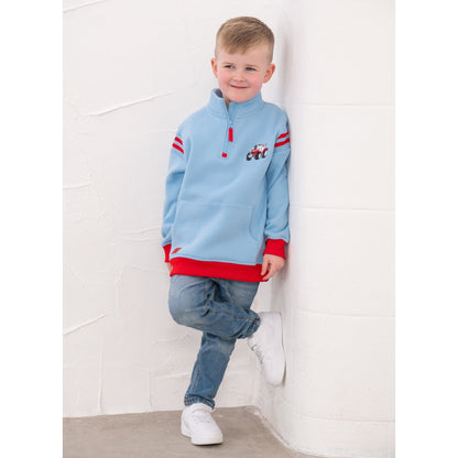 A boy wearing a Lighthouse Jonah 1/4 zip sweatshirt in sky blue with red trim and a red embroidered tractor applique on the chest with blue jeans and white trainers.