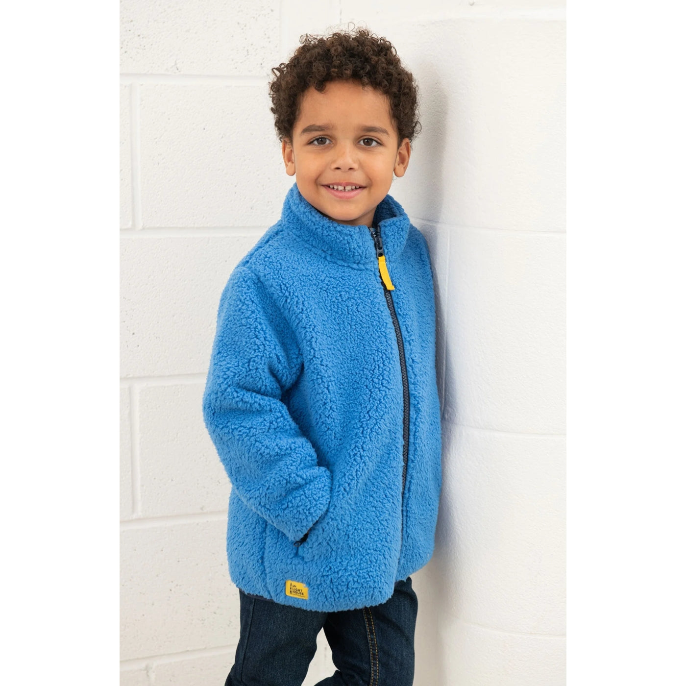 Child wearing a Lighthouse Kian blue fleece jacket with a yellow zipper against a white brick wall.