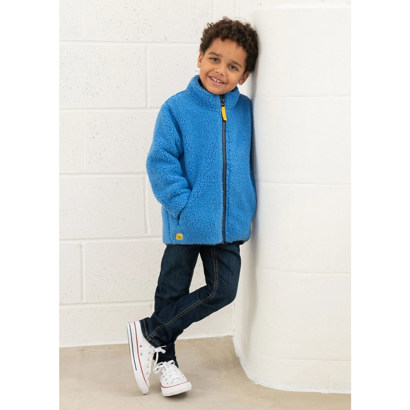 Child wearing a blue Lighthouse Kian sherpa fleece jacket against a white brick wall
