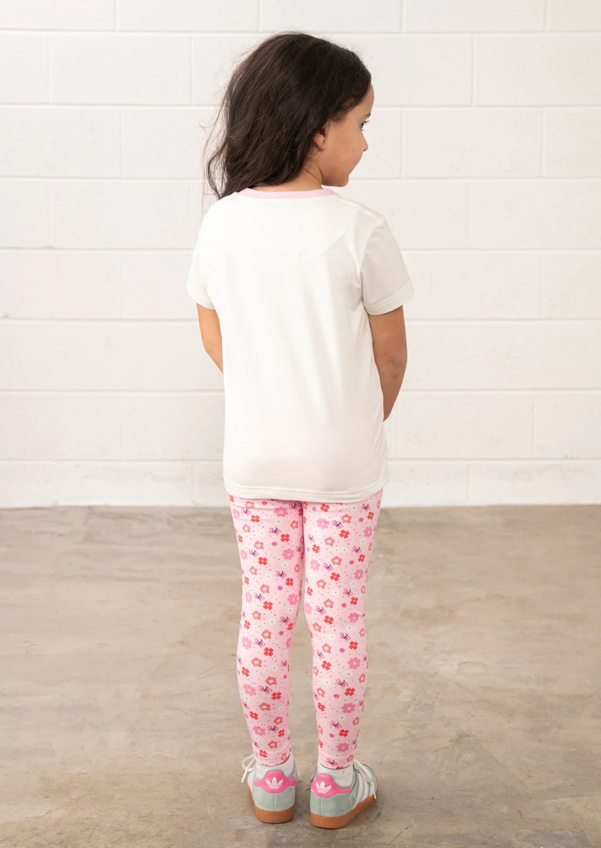 Child wearing Lighthouse Kids pink floral leggings with white t-shirt and sneakers standing against white brick wall