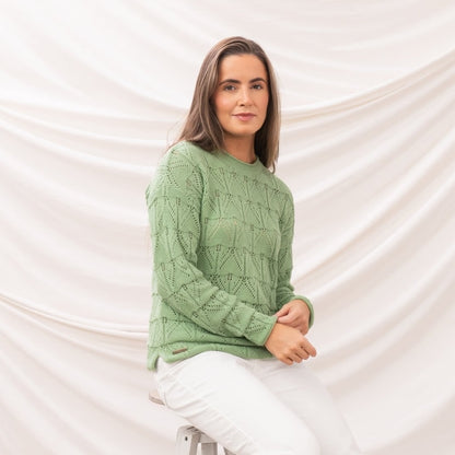 Side view of a woman wearing a Lighthouse Eden knitted jumper in Soft Sage Green with a geometric pointelle knit pattern with white trousers.