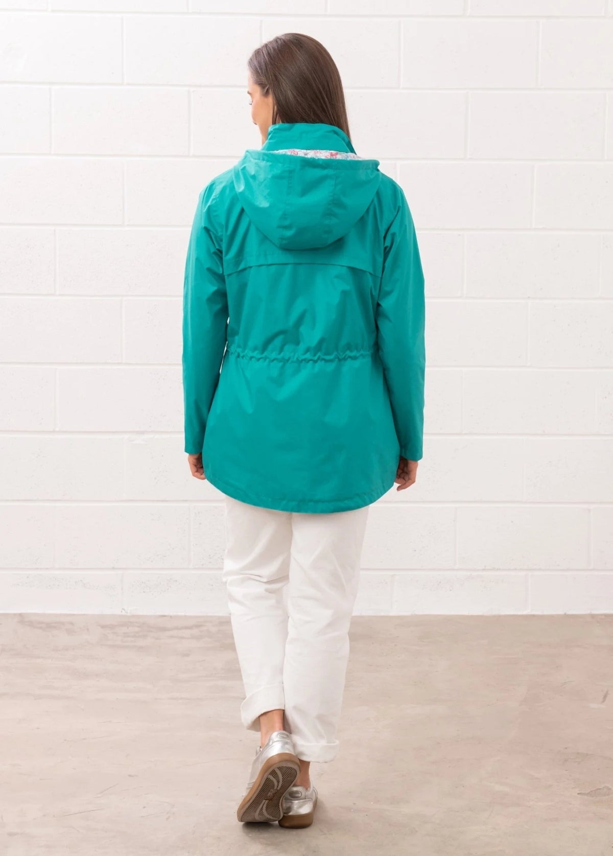 Back view of a woman wearing a Lighthouse teal womens waterproof jacket with hood and elastic waist