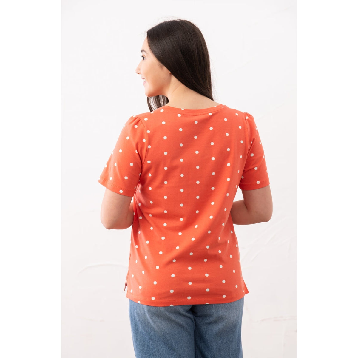 Back view of a woman wearing a Lighthouse short sleeve Ariana v-neck t-shirt in coral with a white dot pattern and blue jeans.