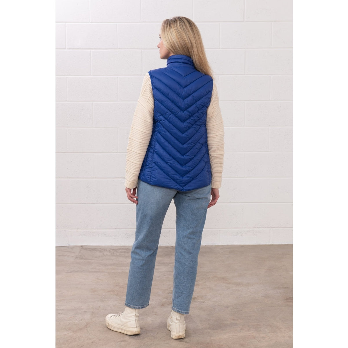 Back view of a woman wearing a Lighthouse padded Laurel Gilet in Indigo Blue over a cream jumper, with blue jeans.