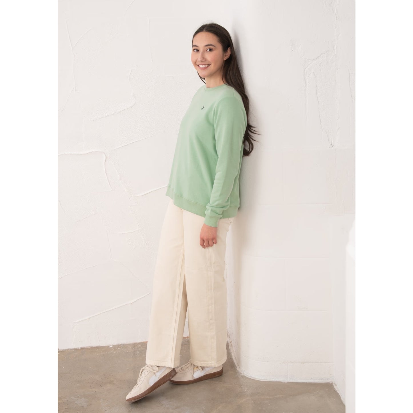 Side view of a woman wearing a Lighthouse Seaside crew neck sweatshirt in Pastel Green with cream trousers.