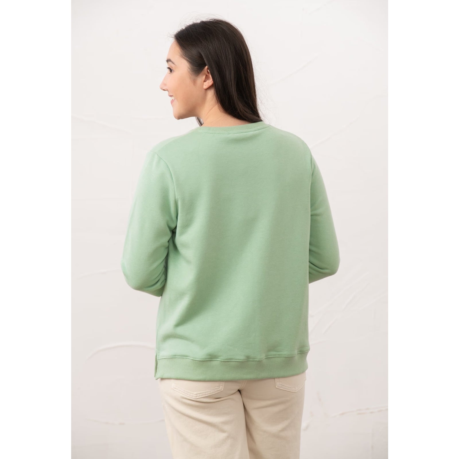 Back view of a woman wearing a Lighthouse Seaside crew neck sweatshirt in Pastel Green with cream trousers.