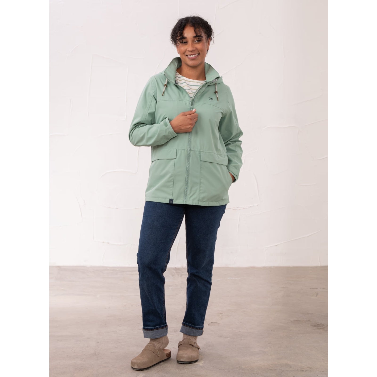 A woman wearing a Lighthouse waterproof Beachcomber II waterproof jacket in Basil green with blue jeans.