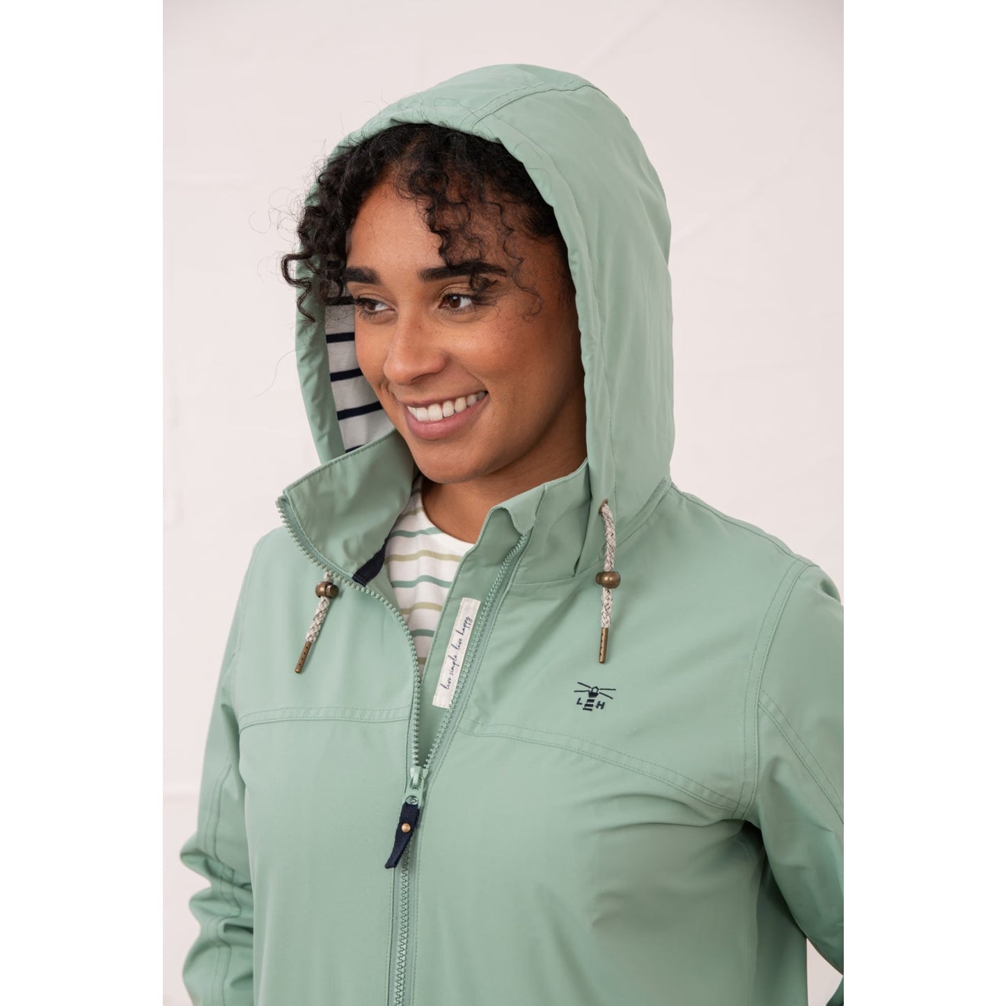 Close-up view of a woman wearing a Lighthouse waterproof Beachcomber II waterproof jacket in Basil green with the hood up.