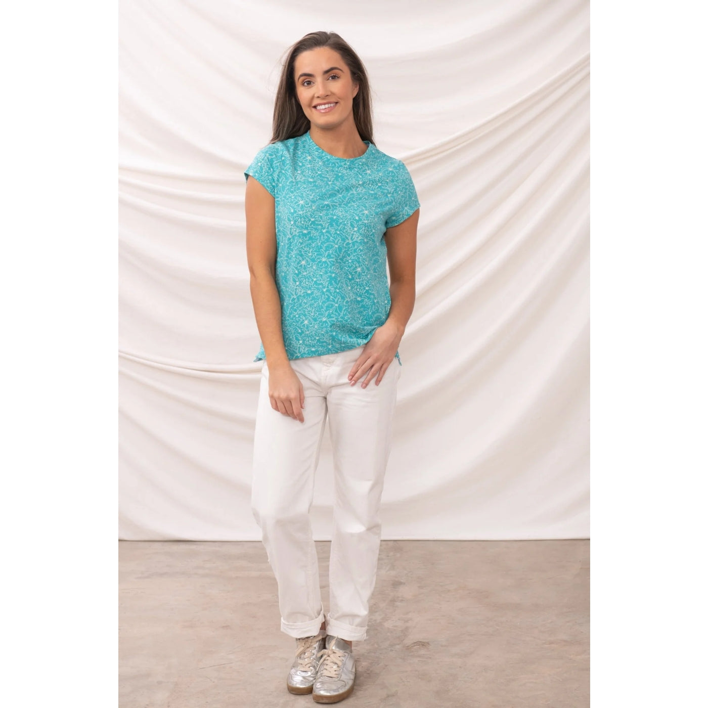 Woman wearing a teal Lighthouse Ccauseway short sleeve t-shirt with a white floral pattern and white pants against a plain background