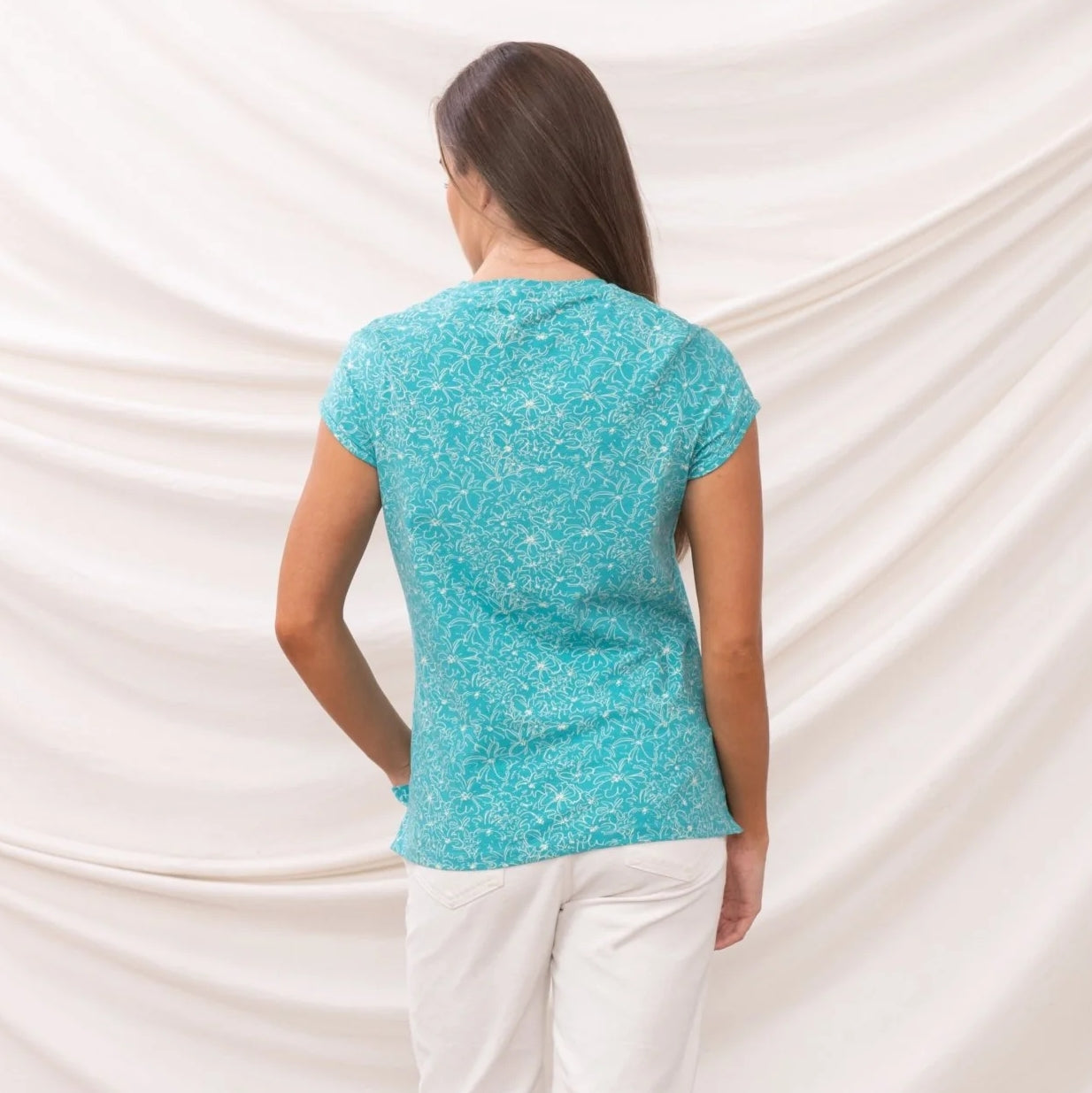 Back view of a woman wearing a Lighthouse Causeway short sleeve tee in Soft Teal with a white floral pattern and white pants against a white background