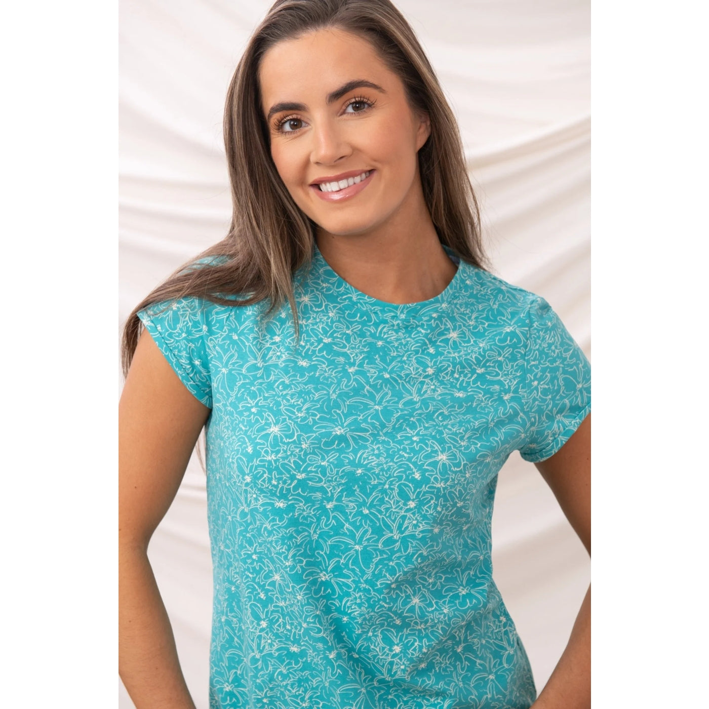Front view of a woman wearing a Lighthouse short sleeve Causeway t-shirt in teal with a floral pattern against a white background