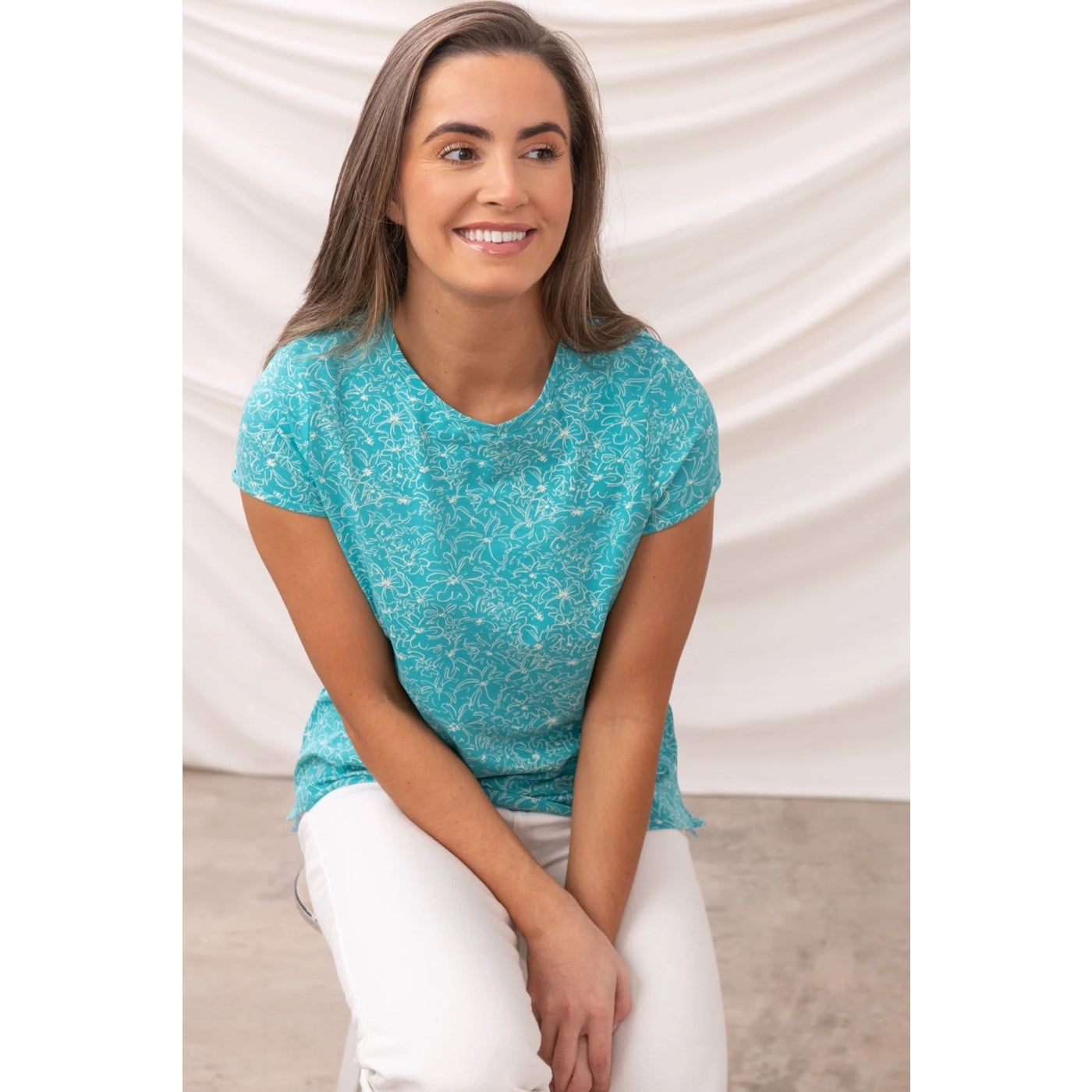 Woman wearing a Lighthouse short sleeve causeway t-shirt in Soft Teal with a floral pattern sitting on a white background