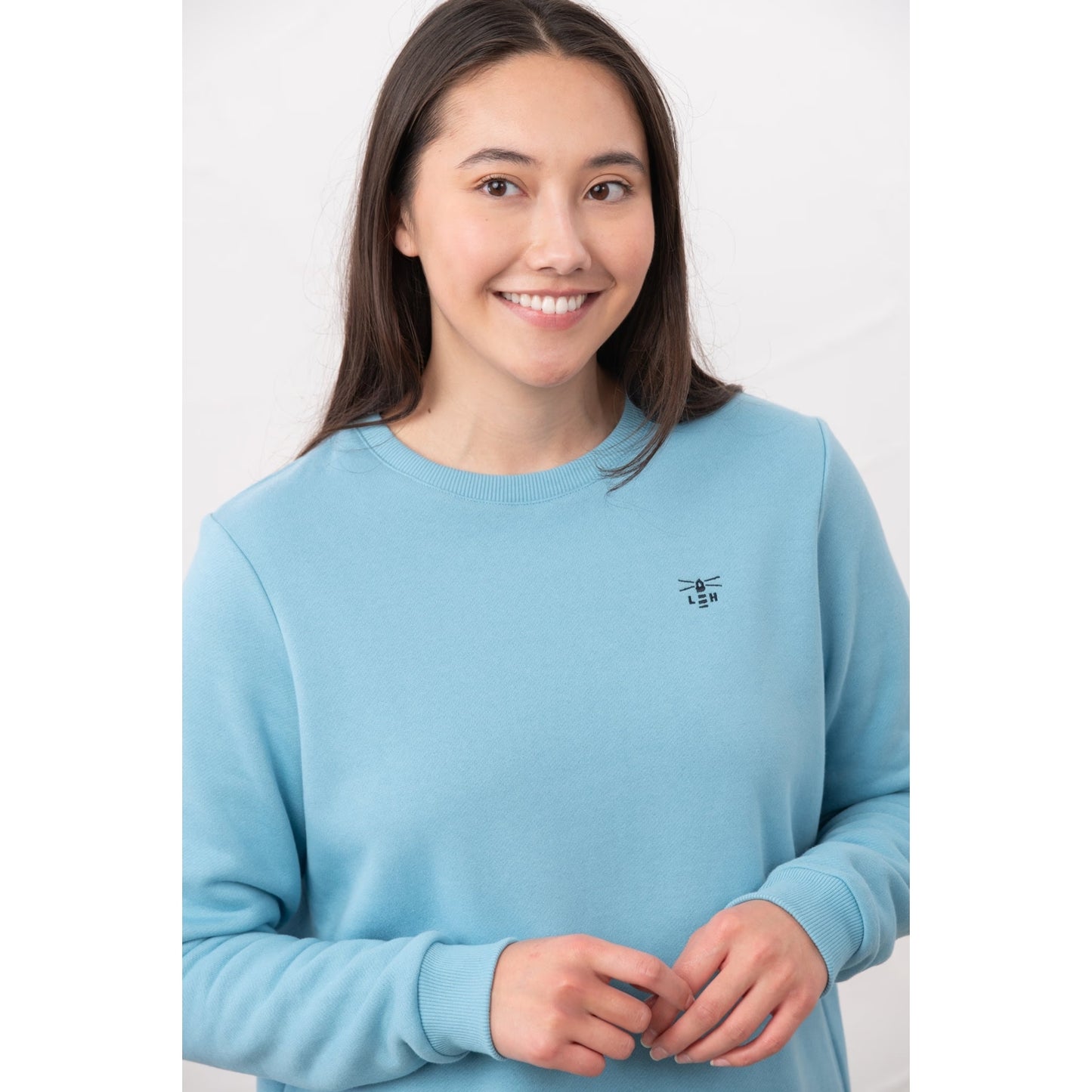 Close-up view of a woman wearing a Lighthouse Seaside crew neck sweatshirt in blue with small logo embroidered on one side of the chest and elasticated cuffs.