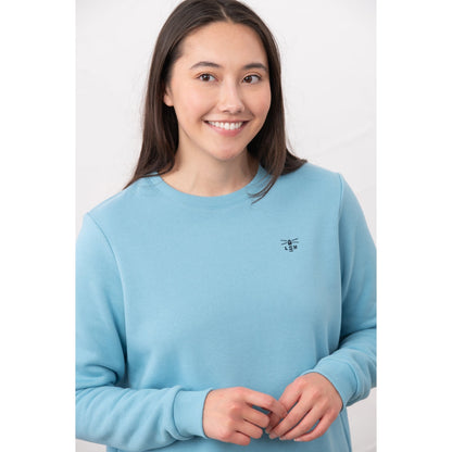 Close-up view of a woman wearing a Lighthouse Seaside crew neck sweatshirt in blue with small logo embroidered on one side of the chest and elasticated cuffs.