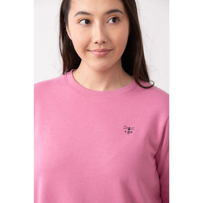 Close-up view of a woman wearing a Lighthouse Seaside crew neck sweatshirt in pink with small logo embroidered on one side of the chest and elasticated cuffs.
