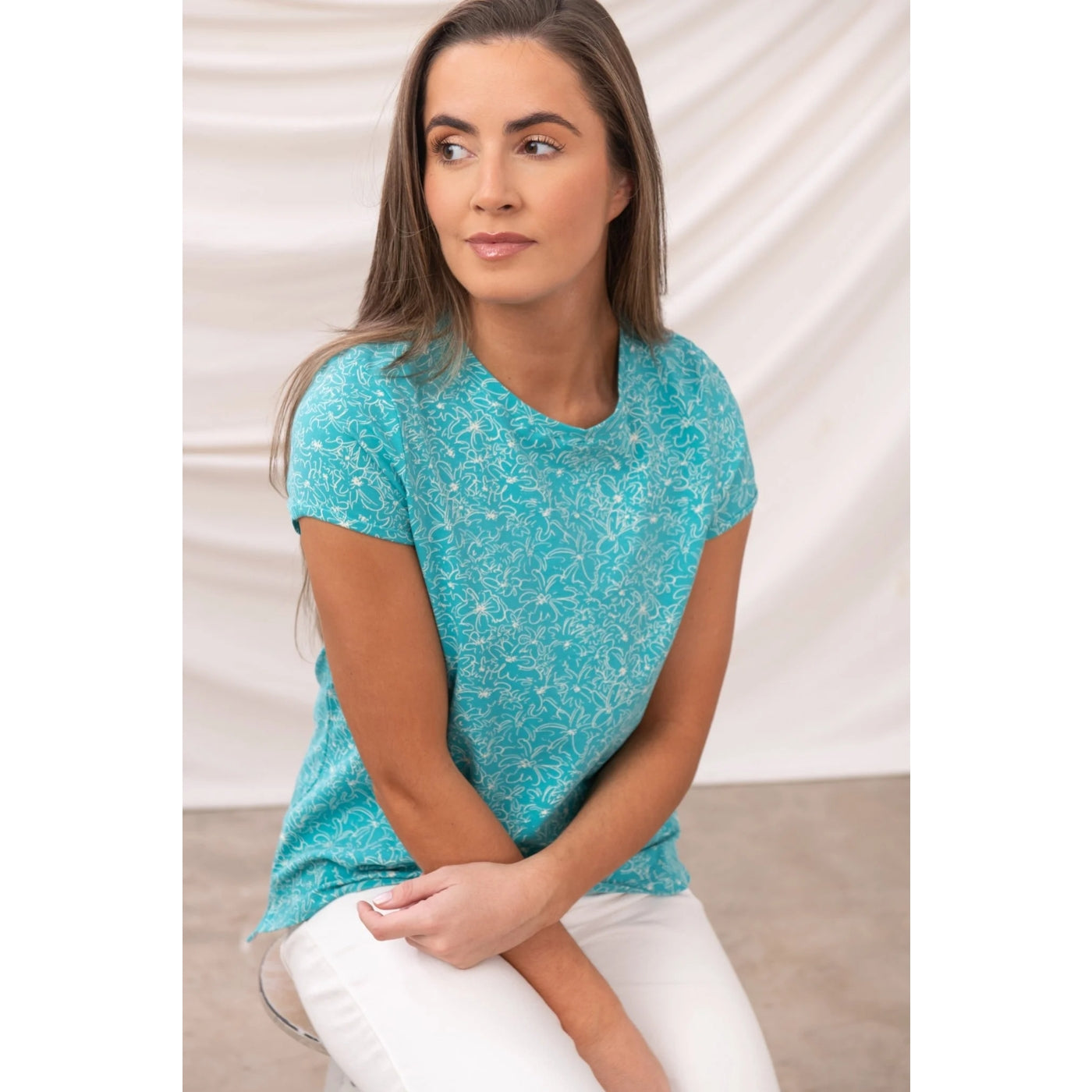Woman wearing a Lighthouse Causeway short sleeve floral pattern t-shirt in Teal sitting on the floor against a white background