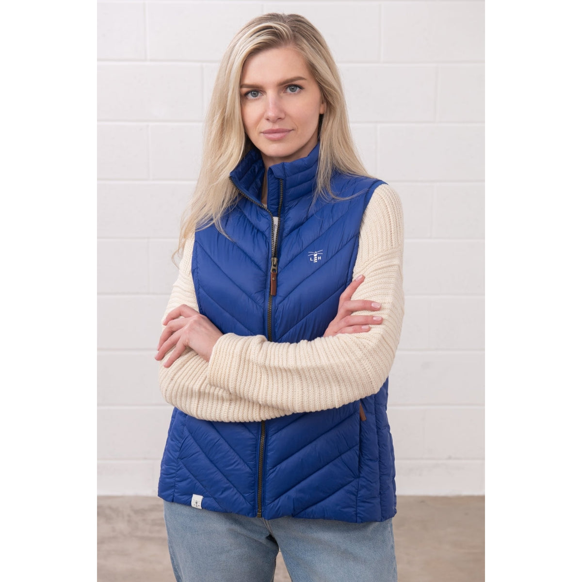 Front view of a woman wearing a Lighthouse padded Laurel Gilet in Indigo Blue over a cream jumper with her arms crossed.