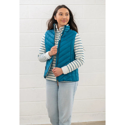 Woman wearing a Lighthouse Laurel puffer gilet in teal blue over a striped shirt against a white brick wall.