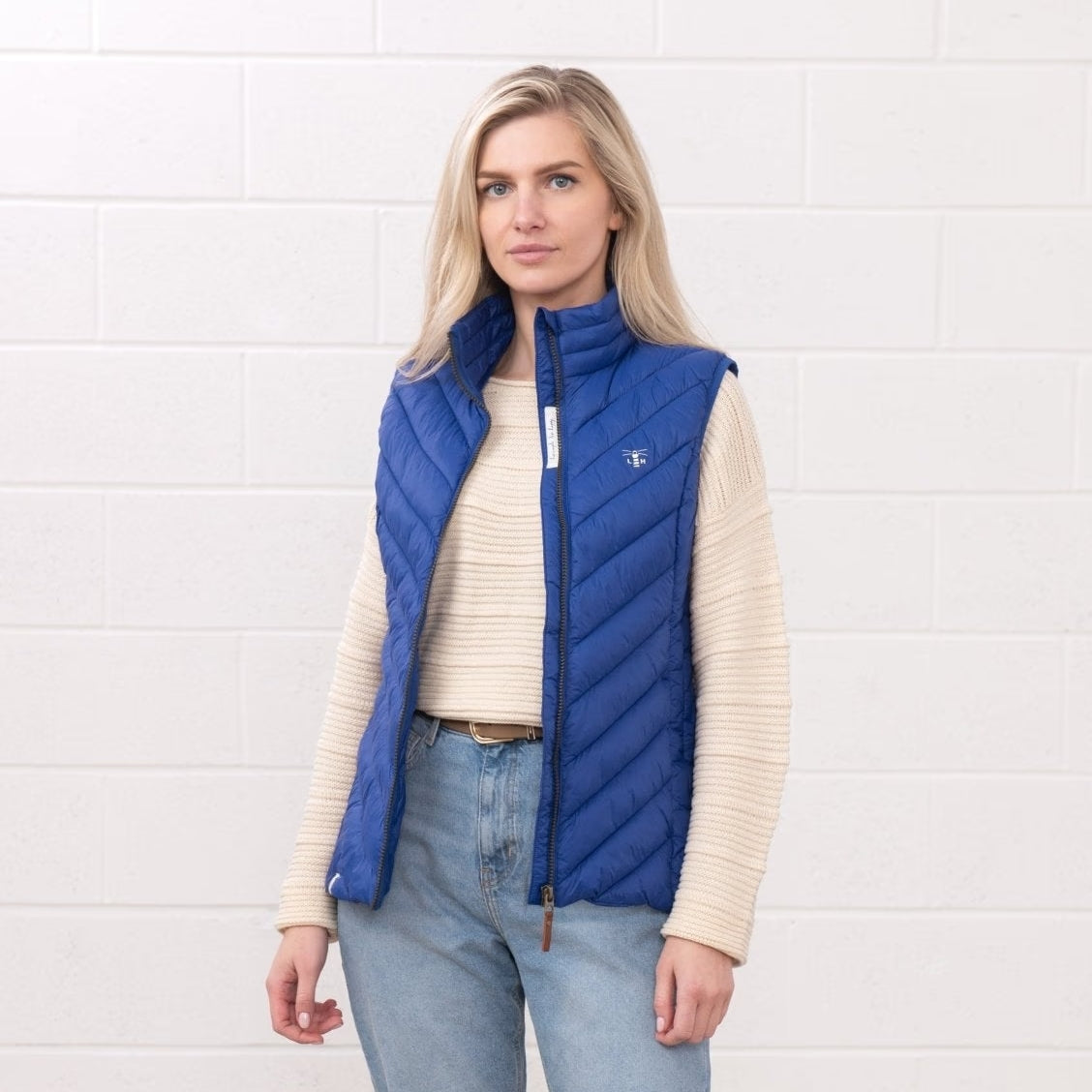 Front view of a woman wearing a Lighthouse padded Laurel Gilet in Indigo Blue unzipped over a cream jumper.