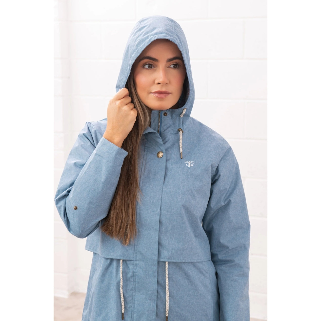 Woman wearing a Lighthouse Pippa 3/4 length waterproof raincoat in Soft Denim Blue with the hood up.