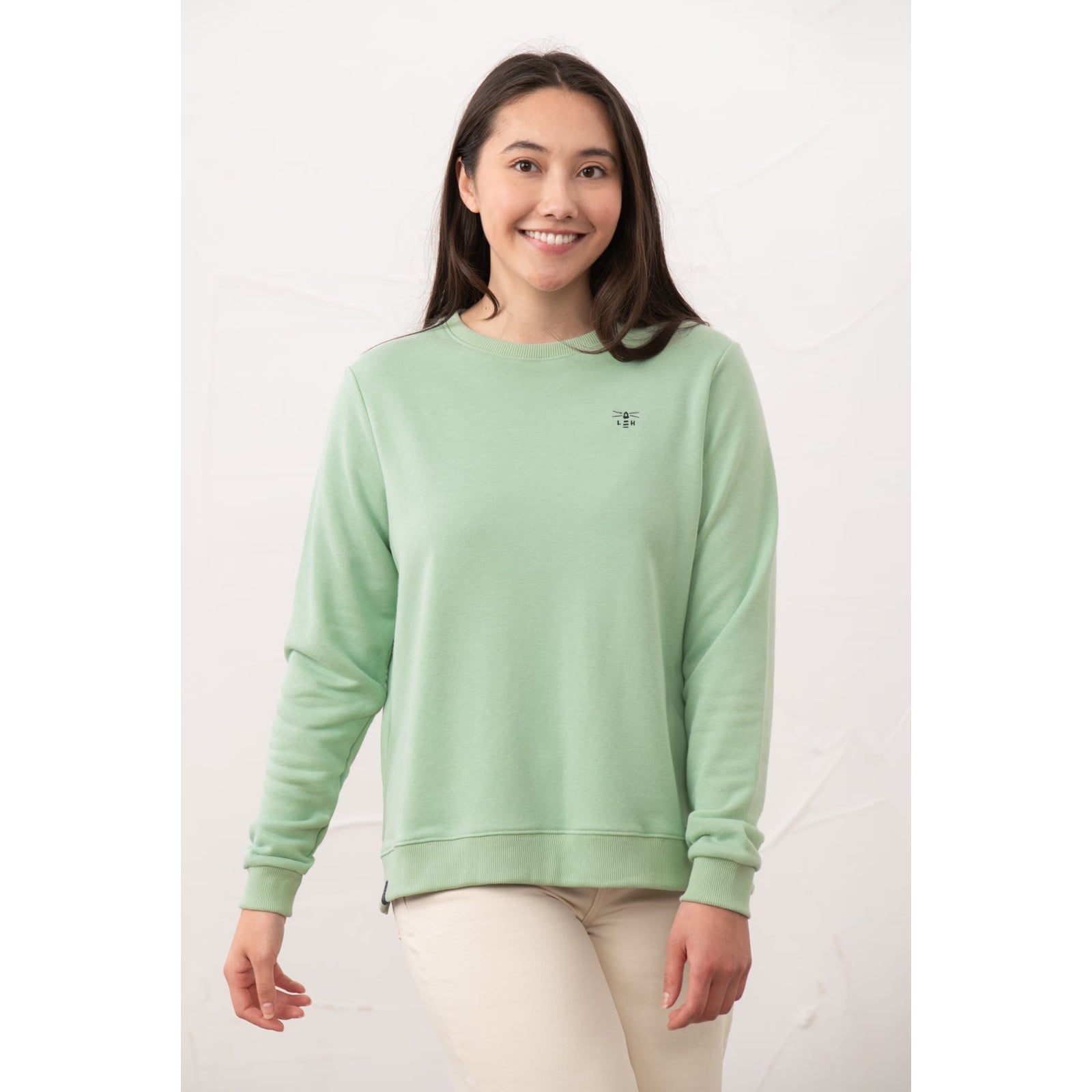 A woman wearing a Lighthouse Seaside crew neck sweatshirt in Pastel Green with small logo on the chest, elasticated cuffs and with cream trousers.