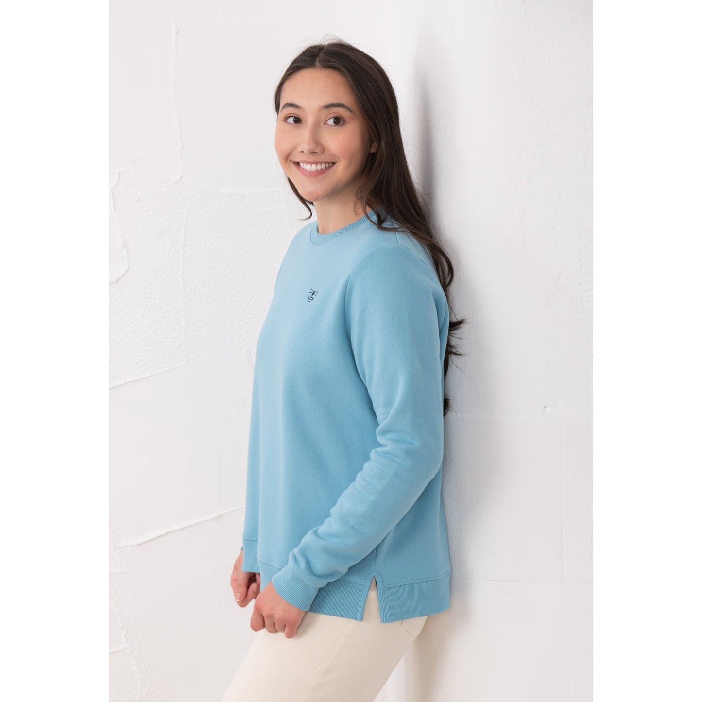 Side view of a woman wearing a Lighthouse Seaside crew neck sweatshirt in blue with small logo embroidered on one side of the chest, elasticated cuffs and side seam slits.