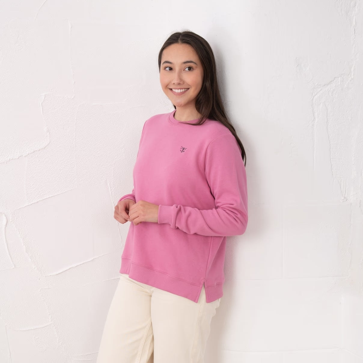 Side view of a woman wearing a Lighthouse Seaside crew neck sweatshirt in pink with small logo embroidered on one side of the chest, elasticated cuffs and side seam slits.