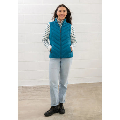 Woman wearing a Lighthouse Laurel puffer gilet in teal blue against a white brick wall.