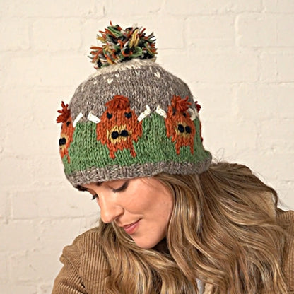 Woman wearing a knitted Pachamama bobble hat with a Highland Cow design against a white brick wall.