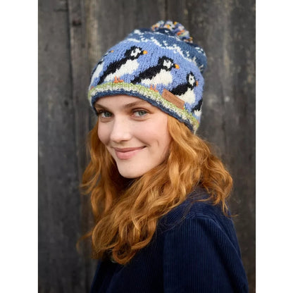 Woman wearing a blue knitted Pachamama knitted hat with puffin design against a wooden background
