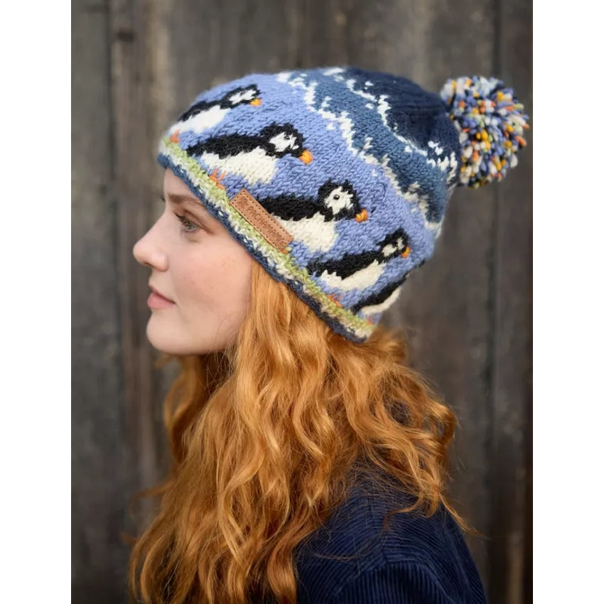 Woman wearing a knitted Pachamama bobble hat with puffin design against a wooden background