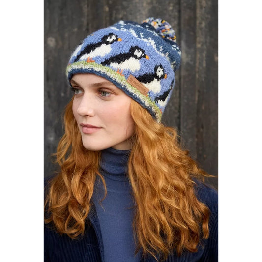 Woman wearing a blue Pachamama knitted hat with puffin design against a wooden background