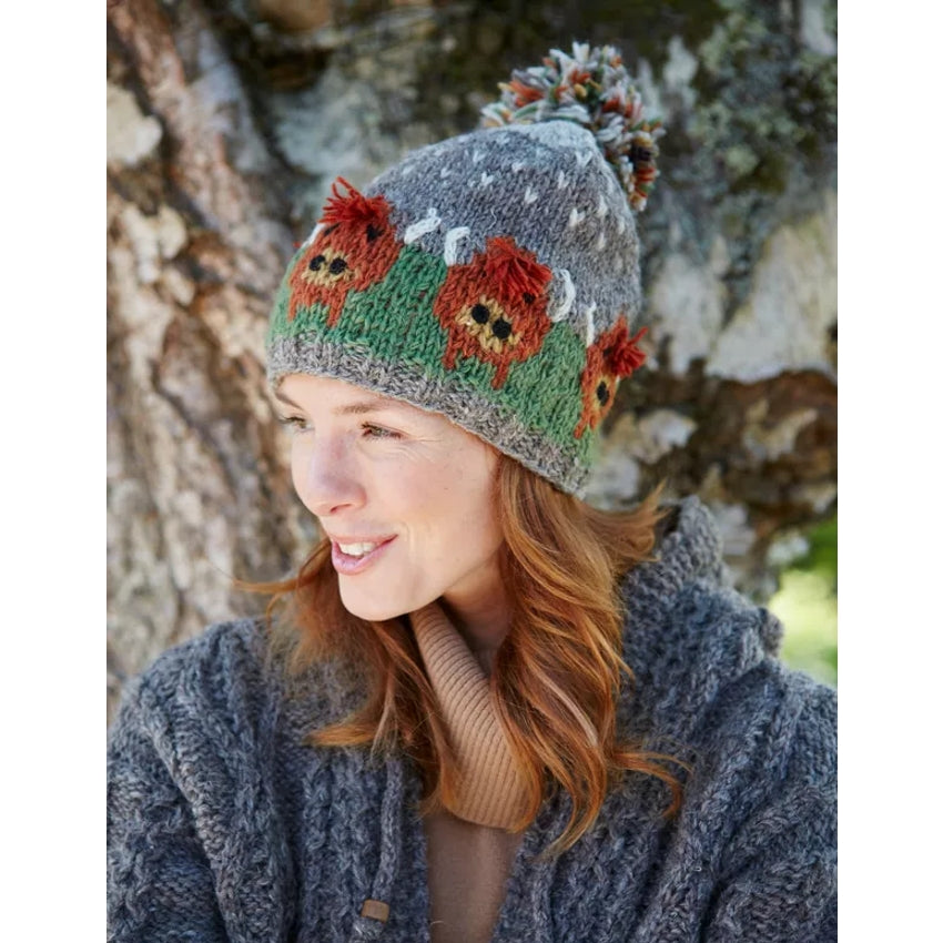 Main view of a woman wearing a Pachamama knitted bobble hat with a Highland Cow design against a natural background
