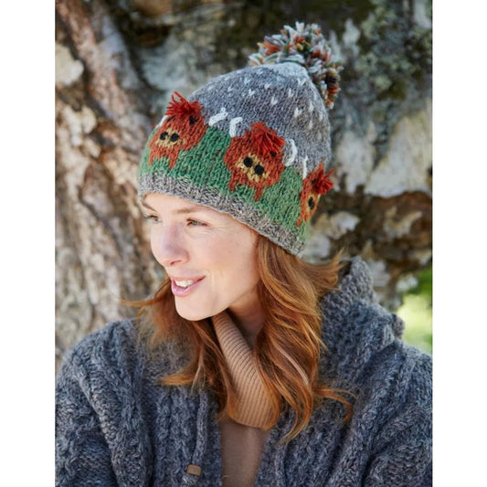 Main view of a woman wearing a Pachamama knitted bobble hat with a Highland Cow design against a natural background