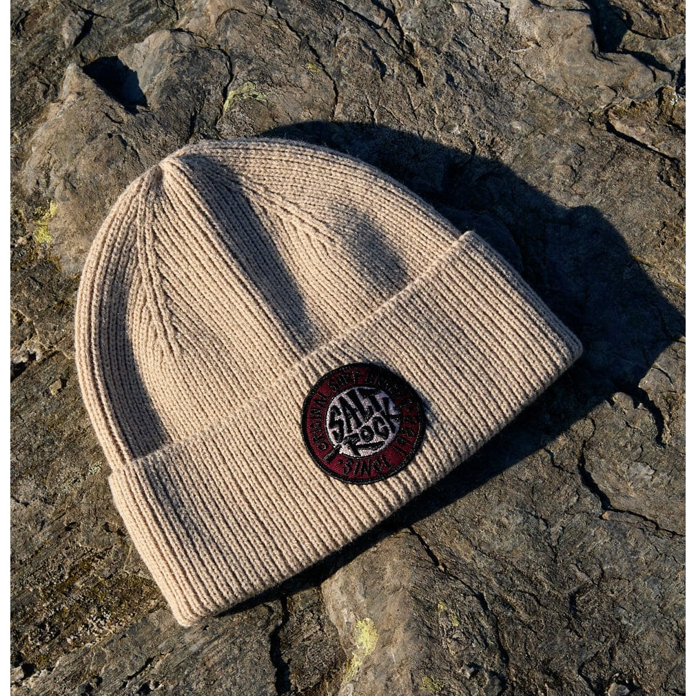 Beige Saltrock knitted beanie with a round logo on a stone surface