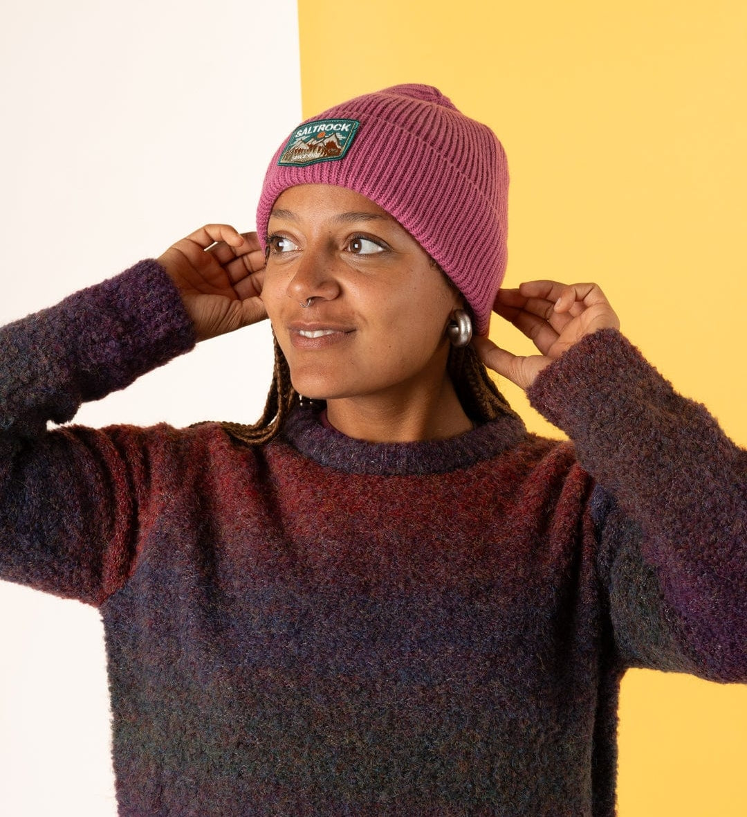 Woman wearing a dark pink Saltrock knitted Uplands beanie with a turn-up hem and mountains logo patch with a  sweater against a yellow background
