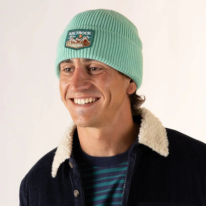 Man wearing a light green Saltrock Uplands beanie with a turn-up hem and mountains logo patch, smiling against a white background