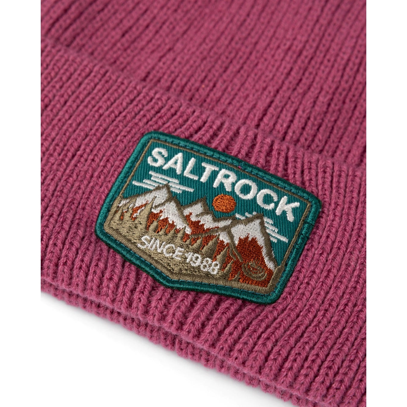 Close up of a dark pink knitted beanie with turn-up hem and Saltrock mountains logo patch on a white background