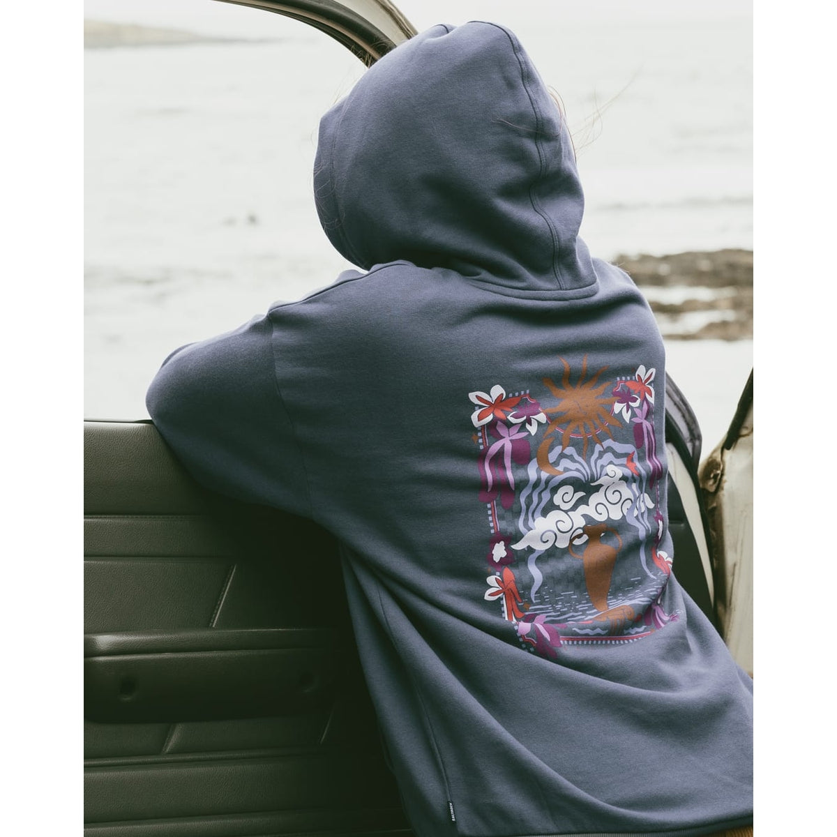 Outdoor back view of a woman wearing a Saltrock Athena pop hoodie in Dark Blue, featuring a large print on the back of a terracotta vase with a coastal themed background.