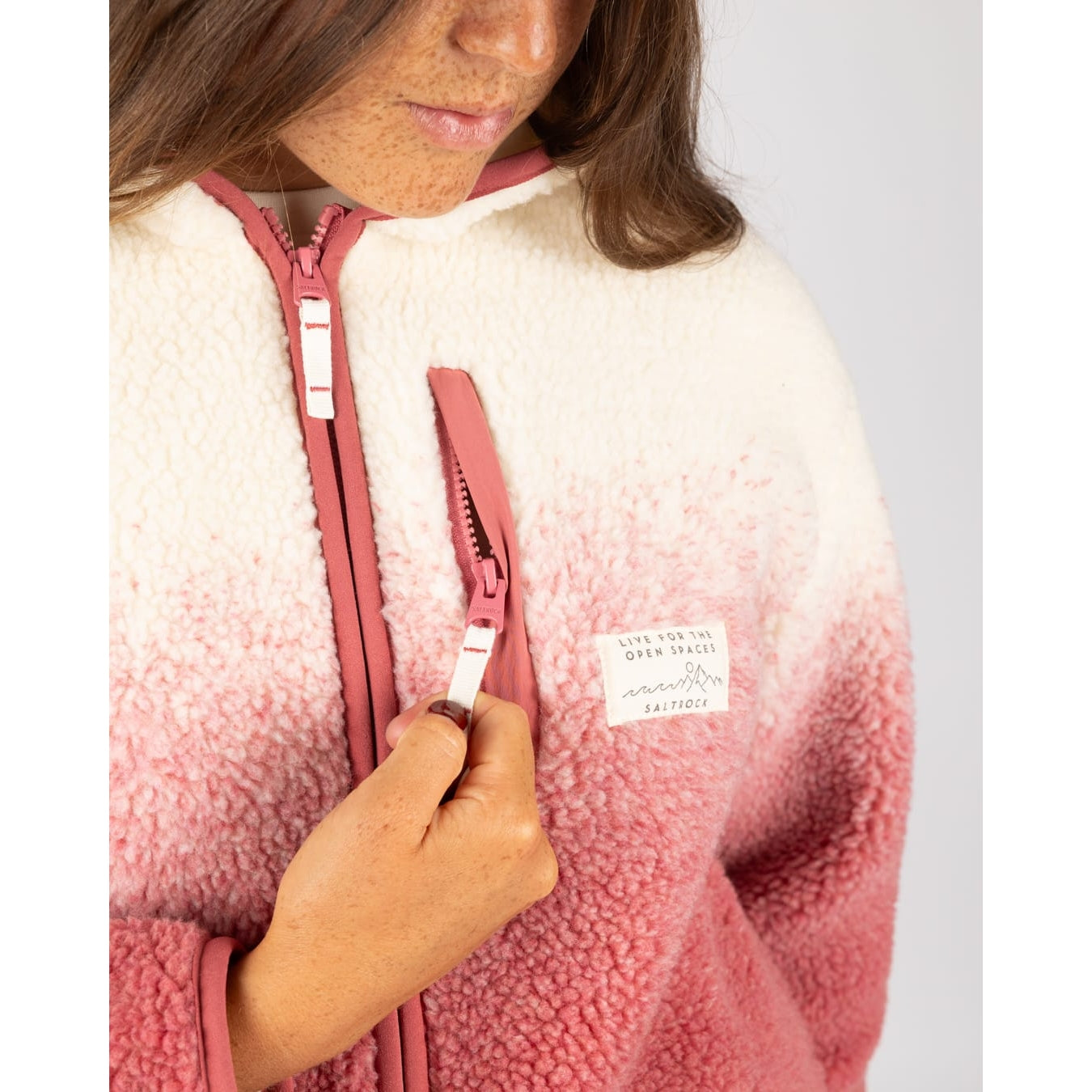 Close-up view of the chest zip pocket of a woman wearing a Saltrock full zip hooded Alba sherpa fleece in a dip dye cream to pink design.
