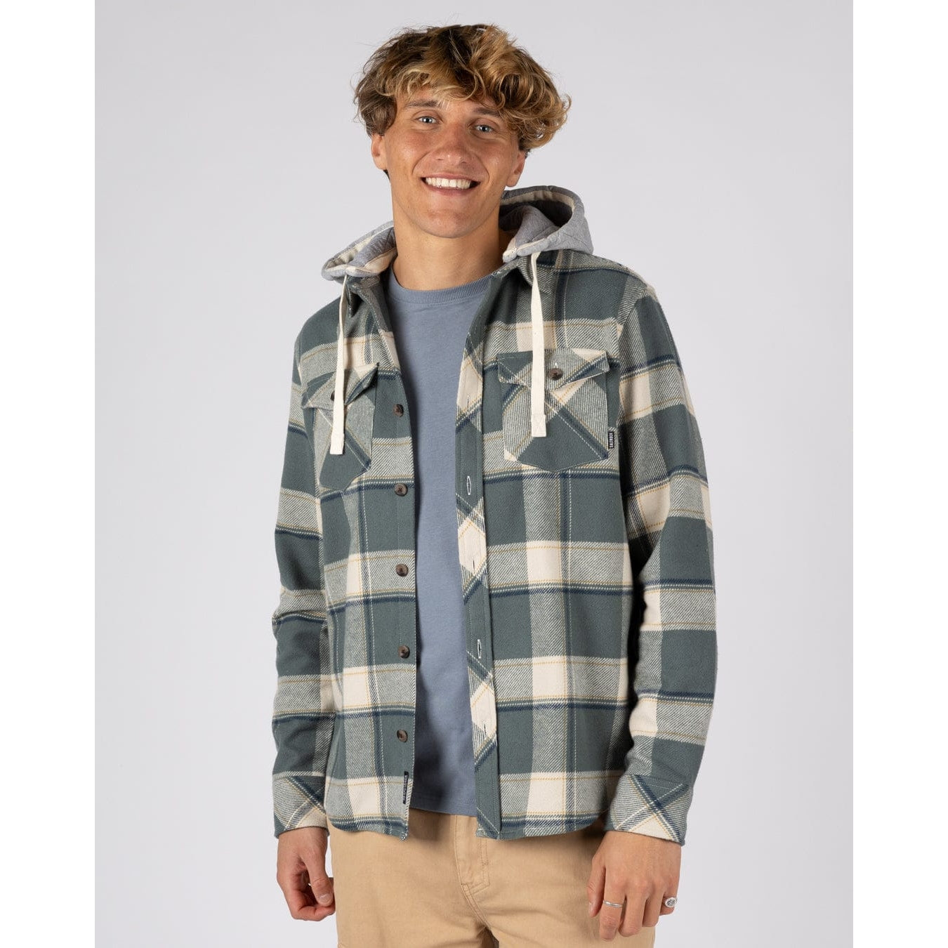 Man wearing a mens Saltrock Strait long sleeve check shirt in dark teal green with chest pockets and a detachable grey hood with tan trousers.