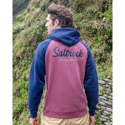 Back view of a man wearing a Saltrock Postcard Script raglan pop hoodie in dark pink with navy blue sleeves and hood and logo print stood outdoors
