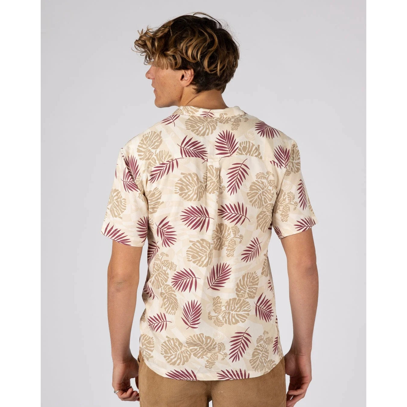 Back view of a man wearing a Saltrock short sleeve Warp Surf Resort shirt in cream with a dark red and tan tropical leaves print with tan shorts