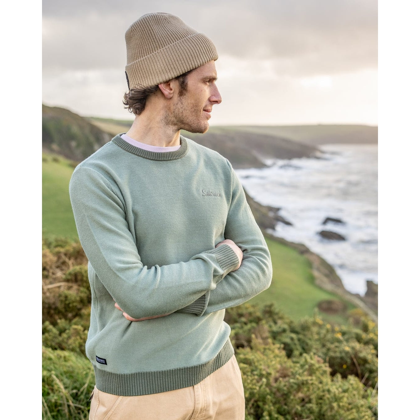 Side view of a man wearing a light green Saltrock Callum knitted jumper standing by a coastal landscape